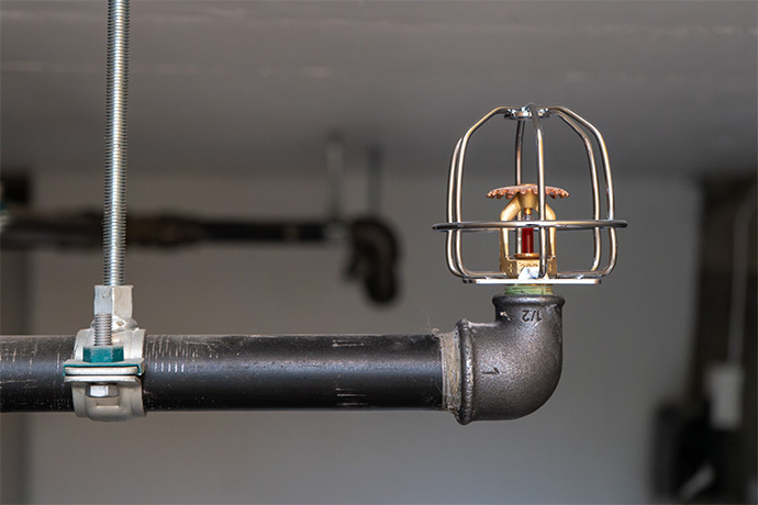Sprinklerhuvud med skyddsbur monterat på svart rörledning i en byggnads tekniska installation.