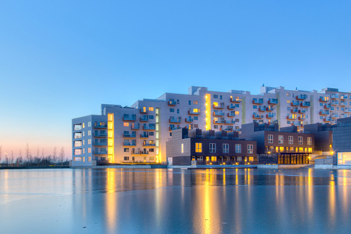 Modern bostadsbyggnader vid vatten i skymning, med upplysta fasader som speglas i en stilla vattenyta.