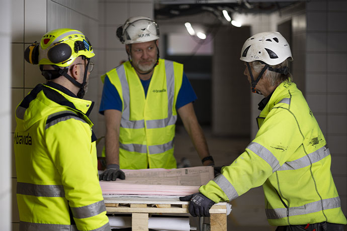 Tre personer i skyddshjälm och varselkläder planerar arbete kring ritningar på en byggarbetsplats.
