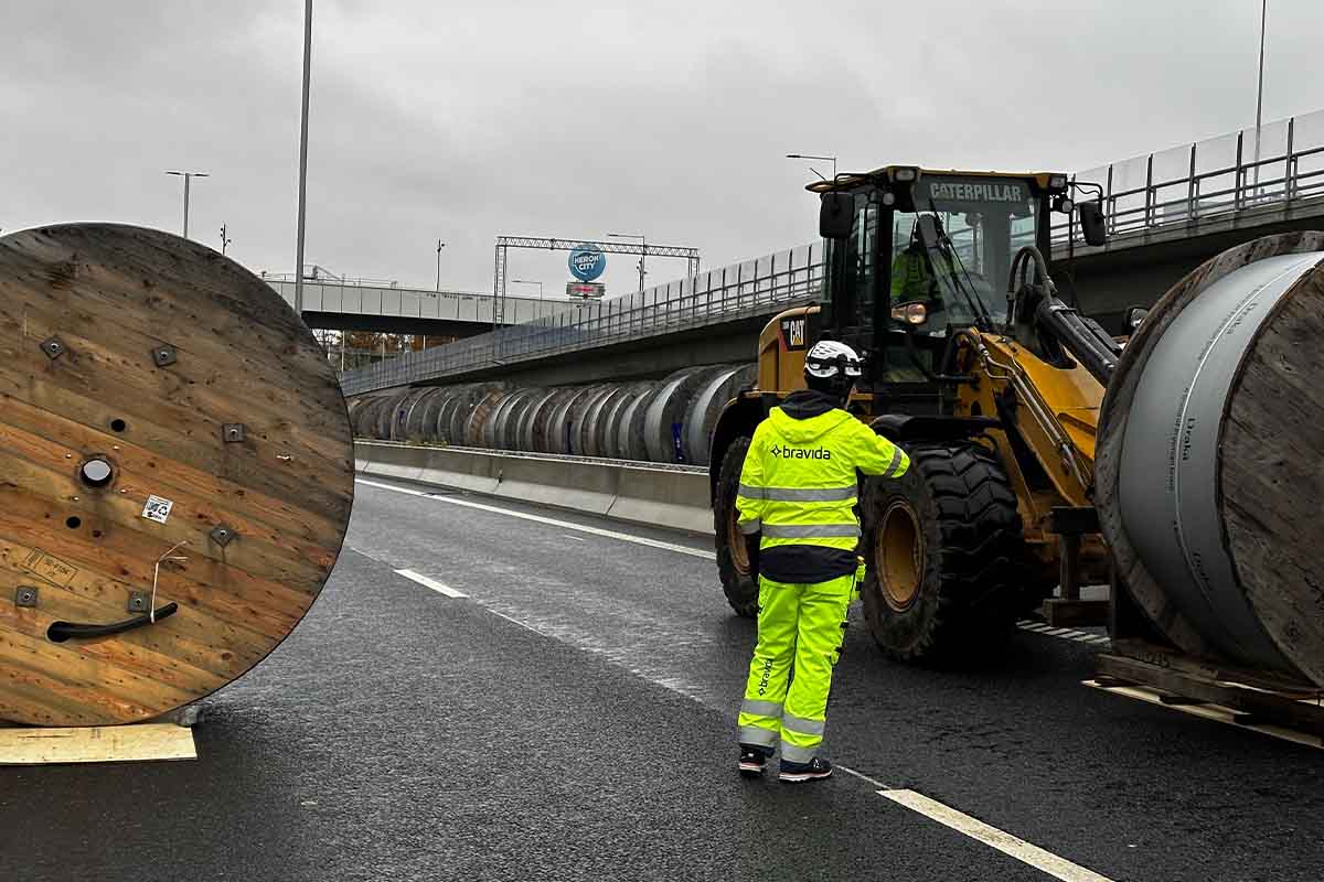 A worker in high-visibility clothing directs a wheel loader transporting large cable drums along a road within the Stockholm Bypass project.