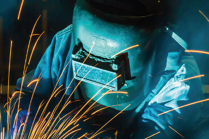 Närbild av svetsarbete i industrimiljö där en person med svetshjälm arbetar med metall.