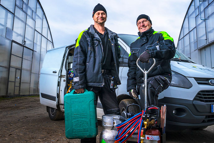 Två kyltekniker står framför en skåpbil med arbetsutrustning utanför ett växthus, redo för tekniskt arbete eller serviceuppdrag. 