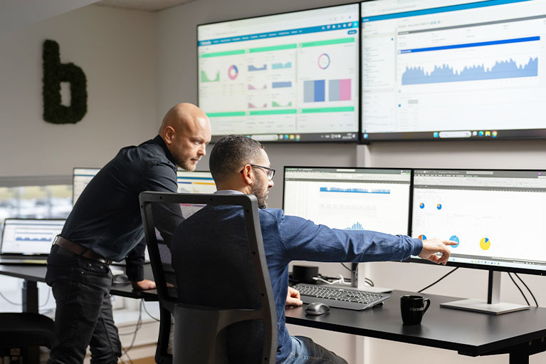 Två personer arbetar vid flera skärmar med data och grafer, analyserar system och följer upp drift i modern kontorsmiljö.