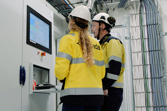 Två personer i Bravidas arbetskläder och skyddshjälmar arbetar vid ett styrskåp med digital skärm i ett teknikutrymme med omfattande el- och kabelinstallationer.